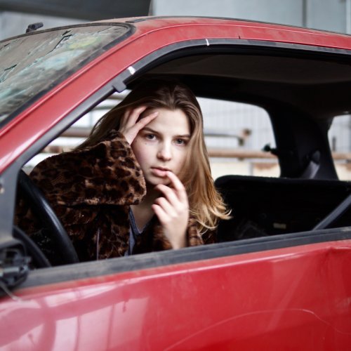 Ein kaputtes Auto mit eingeschlagenen Fenstern in dem ein Model von der Fotografin Felicitas Jander sitzt und ihre Lippen beruehrt.