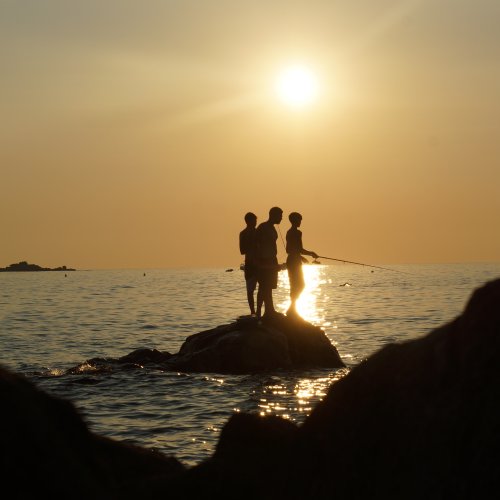 Korsika mit ihrer eleganten Kueste. Drei junge Maenner stehen auf einem Stein und Angeln im Sonnenuntergang.