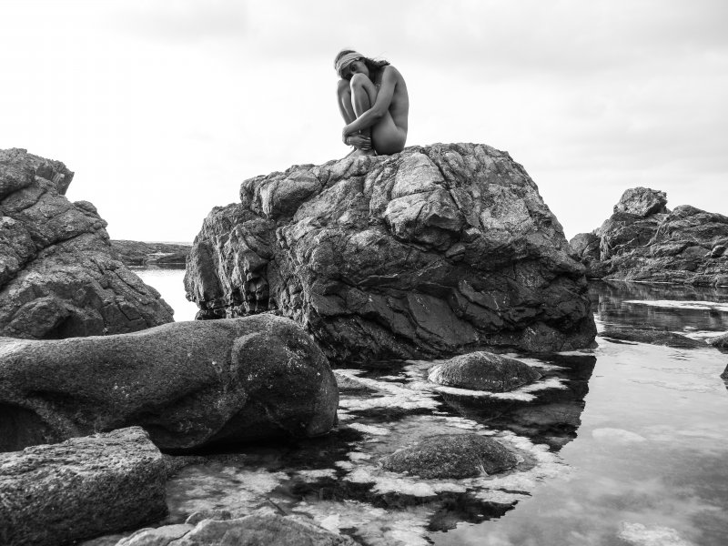 Ein nacktes Model sitzt auf den Felsen der Kueste Korsikas und wird von der Fotografin Felicitas Jander in schwarz-weiss fotografiert.