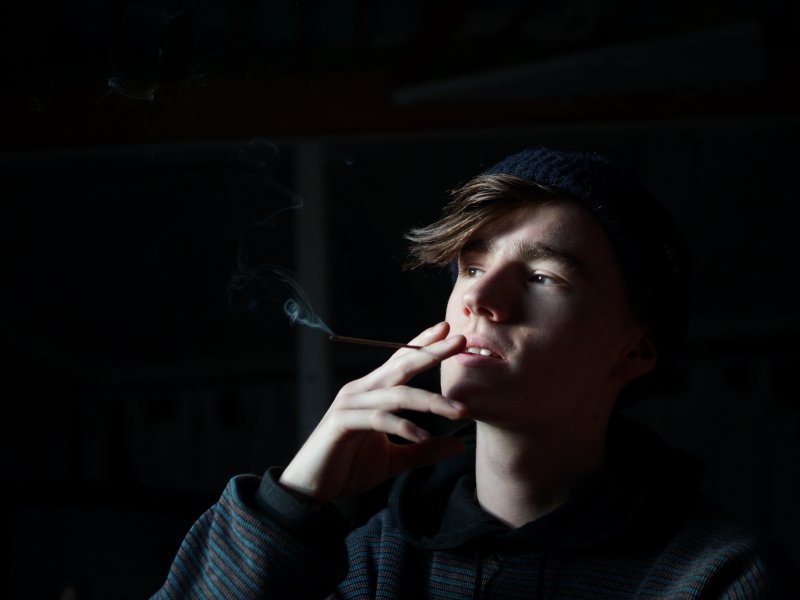 Ein Portrait geschossen von Felicitas Jander, als ein junger Mann ein Raeucherstaebchen wie eine Zigarette verwendet. Er ist schwarz gekleidet.
